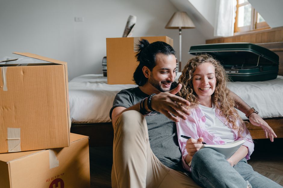A young man and woman sitting on the floor inside a home that is being prepared for a house removal, surrounded by several packed cardboard boxes and a partially open large green suitcase. The woman i