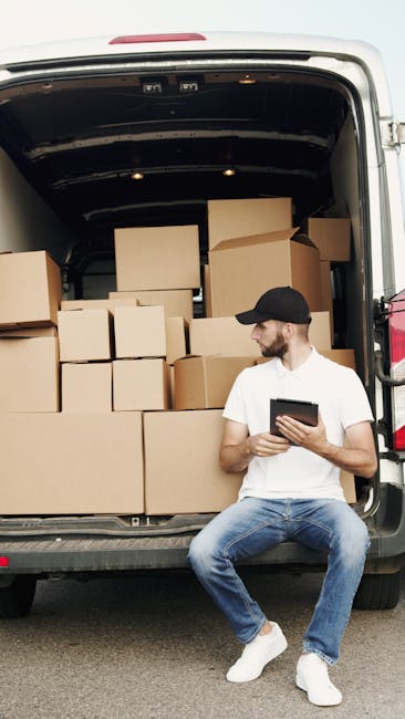 A man wearing a black cap, white T-shirt, and light blue jeans is sitting on the lower bumper of a white moving van with its large rear door open. Inside the van, there are numerous cardboard boxes of