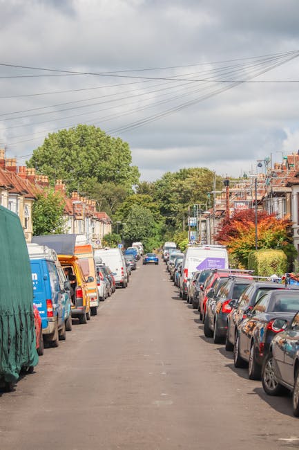 An aerial black-and-white photograph depicts a residential street with lined-up parked cars on both sides of the road, including a white van, a small dark vehicle, and several other cars. The street c