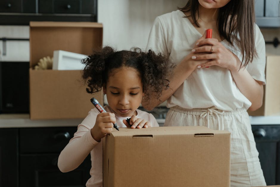 A young child with curly hair is partially visible inside an open cardboard moving box on a wooden floor, with only their head and eyes showing above the box's flaps. Surrounding the child are several